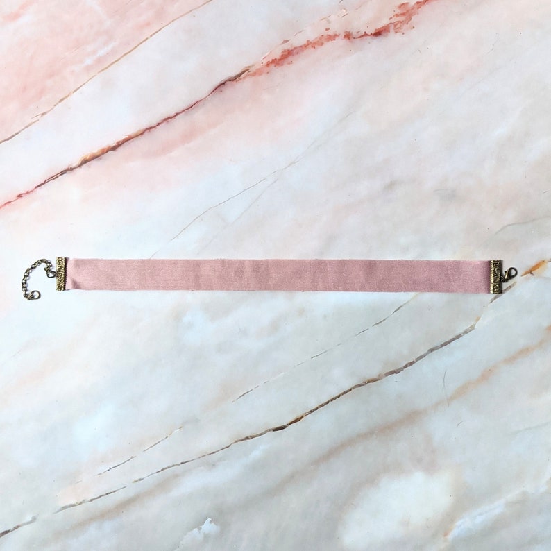 Op de afbeelding: Een delicate roze fluwelen choker met goudkleurige metalen sluitingen. De choker is plat en heeft een eenvoudig ontwerp.