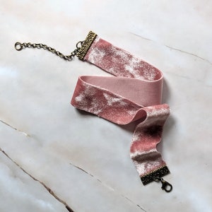 Op de afbeelding: Een roze fluwelen chokerketting met een gouden sluiting. De ketting ligt op een wit marmeren oppervlak.