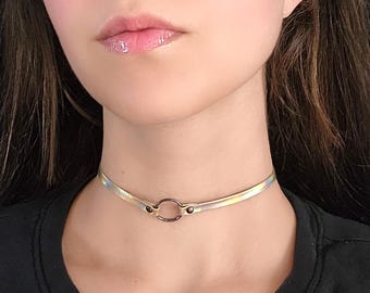 May include: A silver iridescent choker necklace with a black metal ring. The choker is made of a thin, flexible material.