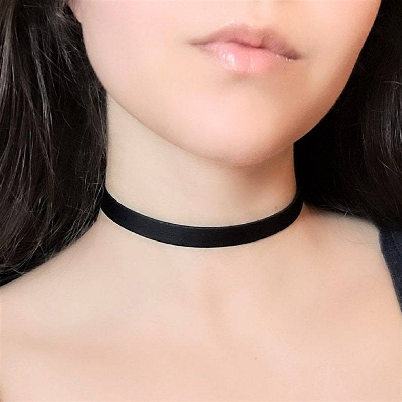 May include: A black choker necklace made of a thin, smooth ribbon. The choker is worn by a person with dark hair.