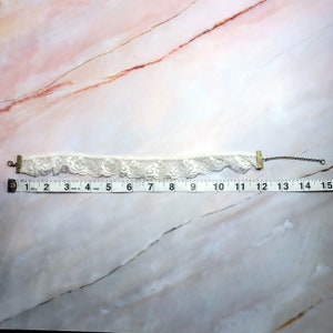 K&ouml;nnte beinhalten: Wei&szlig;e Spitzen-Choker-Halskette mit goldenen Metallverschl&uuml;ssen. Die Halskette ist etwa 30 cm lang.