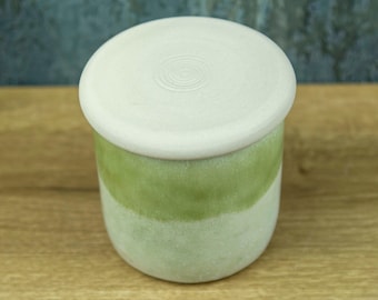 Può includere: Un barattolo cilindrico in ceramica con un coperchio bianco opaco caratterizzato da scanalature circolari concentriche. Il corpo del barattolo presenta uno smalto bicolore, con una fascia verde lime sopra una base verde menta pallido.