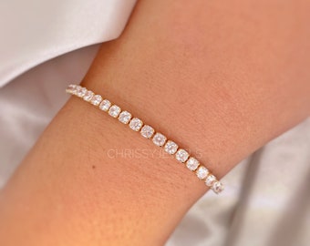Puede incluir: Una pulsera de tenis dorada con piedras preciosas claras de corte cuadrado. La pulsera se lleva en una muñeca, con el nombre de la marca "CHRISSYJEWELS" visible. El fondo es una tela blanca suave.