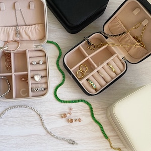 May include: Three different jewelry boxes with compartments for rings, earrings, and necklaces. The boxes are beige, black, and white. The boxes are open and show the compartments inside. There are several pieces of jewelry in the boxes, including a silver chain necklace, a gold chain necklace, a silver bracelet, a gold bracelet, and several rings.