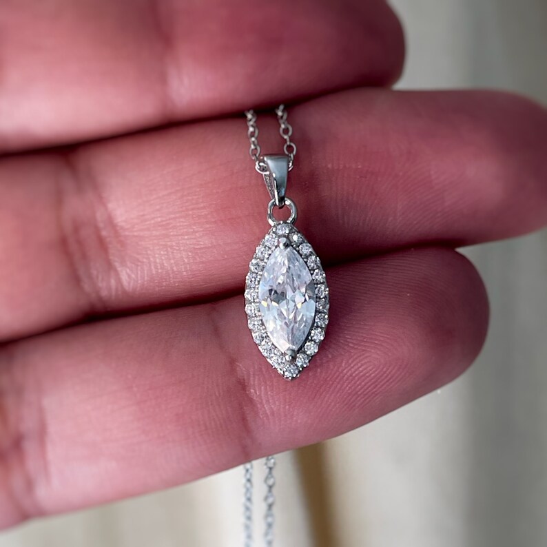 May include: A silver necklace featuring a marquise-cut pendant with a halo of small, clear stones. The pendant is suspended from a delicate chain. The necklace is displayed against a soft, neutral background.
