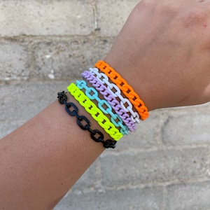 Op de afbeelding: Een verzameling kleurrijke schakelarmbanden in verschillende tinten, waaronder zwart, neongeel, lichtblauw, lavendel, wit en oranje. De armbanden worden gestapeld op een pols gedragen, waarbij hun levendige kleuren en gedurfde ontwerp worden getoond.