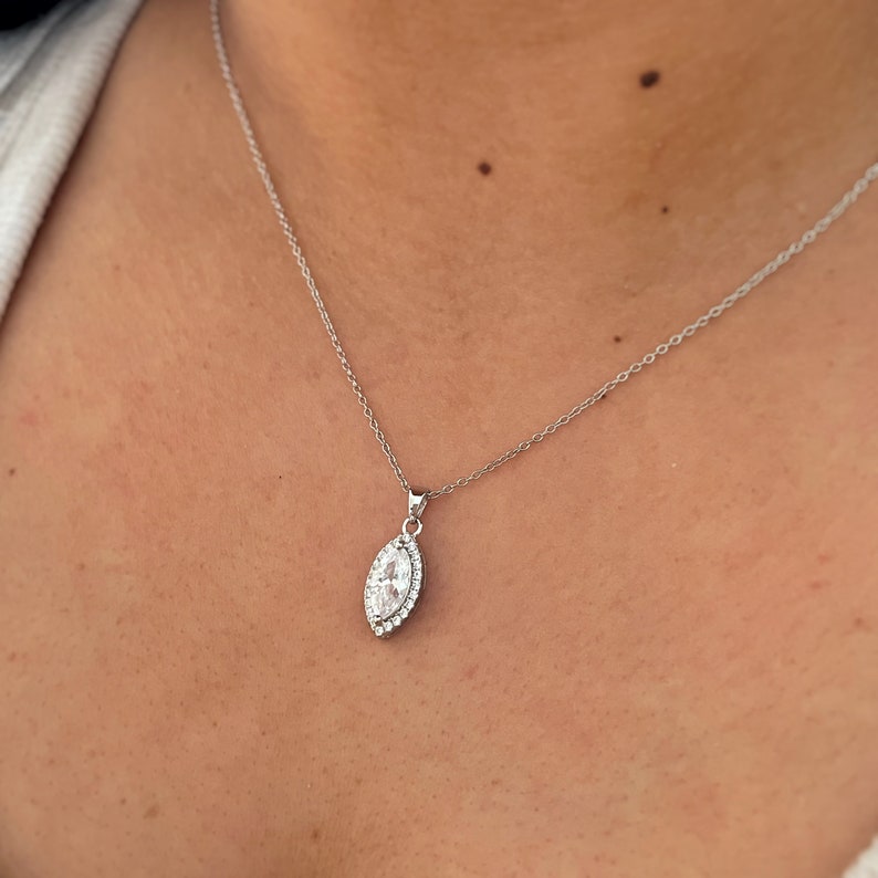 May include: A silver necklace with a pendant featuring a marquise-shaped clear stone surrounded by smaller clear stones. The pendant hangs from a delicate silver chain, creating a classic and elegant look. The necklace is displayed against a skin tone background.