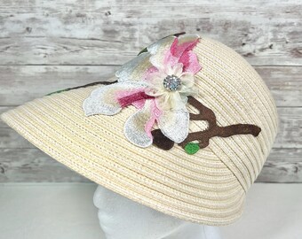 May include: A cream-colored sun hat with a wide brim, adorned with a floral applique. The applique features a pink and white flower with a sparkling center, set on a brown branch with green leaves. The hat is displayed on a white mannequin head.