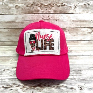 May include: Pink baseball cap with a white patch that says "Nurse Life" with a cartoon image of a woman wearing a face mask and stethoscope.