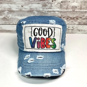May include: A distressed blue denim military-style cap featuring a rectangular white fabric patch on the front. The patch displays the black text "GOOD" above the multicolored, rainbow-gradient text "VIBES" enclosed in black curly brackets.