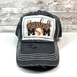 May include: A charcoal gray distressed baseball cap featuring a rectangular white patch on the front. The patch displays the text Baseball MOM in black script and block lettering, with a leopard print background and a baseball graphic.