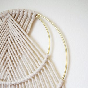 以下が含まれることがあります： 金色の金属製の輪が付いた白いマクラメの壁掛け。マクラメは、アーチ状のシリーズを作る結び目の技術で作られています。