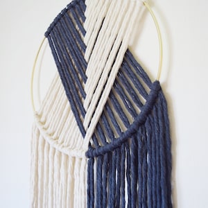May include: A white and navy blue macrame wall hanging with a gold metal hoop. The hanging features a geometric pattern and long fringe.