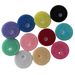Puede incluir: Una colección de once tapas circulares para vasos en varios colores, incluyendo negro, azul, amarillo, morado, verde, rosa, rojo, turquesa, blanco y naranja. Cada tapa está cubierta de pequeños tachones facetados y tiene un orificio central.