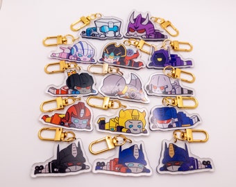 May include: A set of 15 colorful cartoon character keychains featuring Transformers characters. The keychains are made of clear acrylic with printed images of the characters. The keychains are attached to gold-colored metal rings.