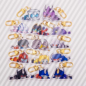 May include: A collection of fifteen clear acrylic keychains featuring colorful, stylized cartoon robot characters in various poses. Each keychain is equipped with a gold-toned metal lobster clasp for attachment, showcasing diverse designs including red, blue, yellow, purple, and grey robotic figures.