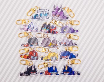 May include: A collection of fifteen clear acrylic keychains featuring colorful, stylized cartoon robot characters in various poses. Each keychain is equipped with a gold-toned metal lobster clasp for attachment, showcasing diverse designs including red, blue, yellow, purple, and grey robotic figures.