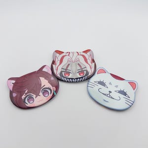 Puede incluir: Tres objetos decorativos con forma de gato con caras de dibujos animados. Uno tiene una cara blanca con una sonrisa astuta, otro tiene un diseño rojo y blanco, y el tercero tiene un personaje de estilo anime con cabello castaño.