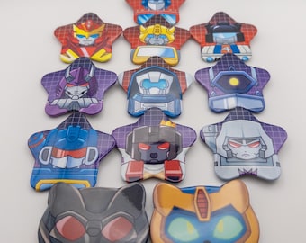 May include: A set of 12 star-shaped pins featuring various Transformers characters. The pins are colorful and feature a variety of characters, including Optimus Prime, Megatron, Starscream, and Bumblebee.