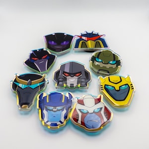 May include: A collection of nine acrylic pins featuring stylized robot head illustrations. The characters display various colors including yellow, blue, green, black, and red with distinct facial features, sharp angles, and metallic-looking outlines.