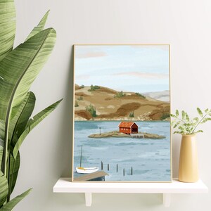 May include: A framed illustration of a red cabin on a small island in a lake. The cabin has a white door and a white roof. The lake is surrounded by green hills. A small white sailboat is docked at a pier in the foreground.