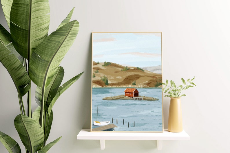 May include: A framed illustration of a red cabin on a small island in a lake. The cabin has a white door and a white roof. The lake is surrounded by green hills. A small white sailboat is docked at a pier in the foreground.
