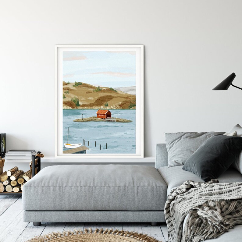 May include: A framed print of a red house on a small island in a blue body of water. The house is surrounded by green hills and trees. The water is calm and reflects the sky.