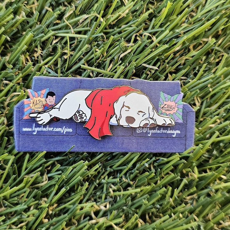 May include: A pin depicting a white dog in a red cape, resting on a blue sofa. The pin incorporates comic book speech bubbles with the words "ZING!" and "KA-POW!". The pin also includes the text "www.lynnfactor.com/pins" and "@lynnfactordesigns".