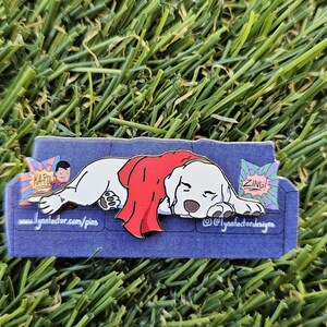 May include: A pin depicting a white dog with a red cape, relaxing on a blue sofa. The pin includes comic book style speech bubbles with the words "ZING!" and "KAPOW!" The pin is set against a background of green grass.