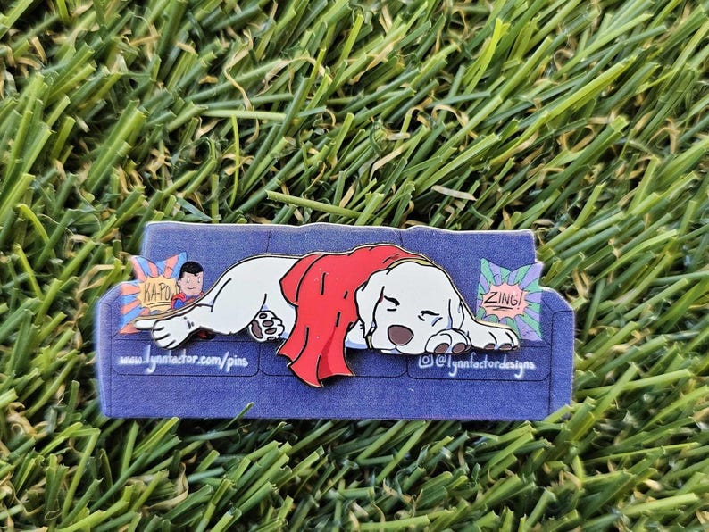 May include: A pin depicting a white dog with a red cape, relaxing on a blue sofa. The pin includes comic book style speech bubbles with the words "ZING!" and "KAPOW!" The pin is set against a background of green grass.