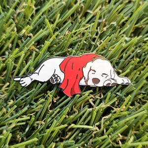May include: A white dog pin with a red blanket, lying down. The dog has brown ears and paws, resting on green grass. This whimsical pin is a charming accessory. The pin is a cute and whimsical accessory.