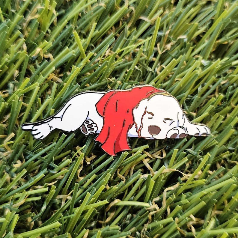 May include: A white dog pin with a red blanket, lying down. The dog has brown ears and paws, resting on green grass. This whimsical pin is a charming accessory. The pin is a cute and whimsical accessory.