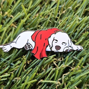 May include: A white dog enamel pin, lying down and wearing a red cape. The dog has brown ears and paws, and is resting on a bed of green grass. The pin is a cute and whimsical accessory.