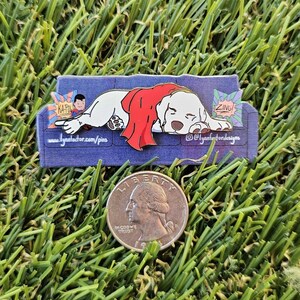May include: A pin depicting a white dog in a red cape, resting on a blue sofa. The pin includes comic book sound effects and the text "www.lynnfordesigns.com/pins" and "@lynnfordesigns".