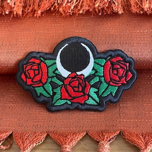 May include: Embroidered patch featuring a black crescent moon and three red roses with green leaves. The patch is on a rust-colored fabric with orange tassels.