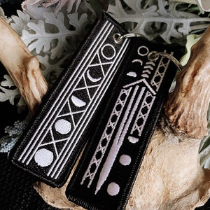 Puede incluir: Dos llaveros de tela negra con bordados blancos. Un llavero presenta un diseño de luna creciente y círculo, el otro presenta un diseño de espada.