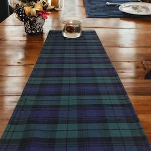 May include: A blue, green, and black plaid table runner. The runner is on a wooden table with a candle in a glass jar.