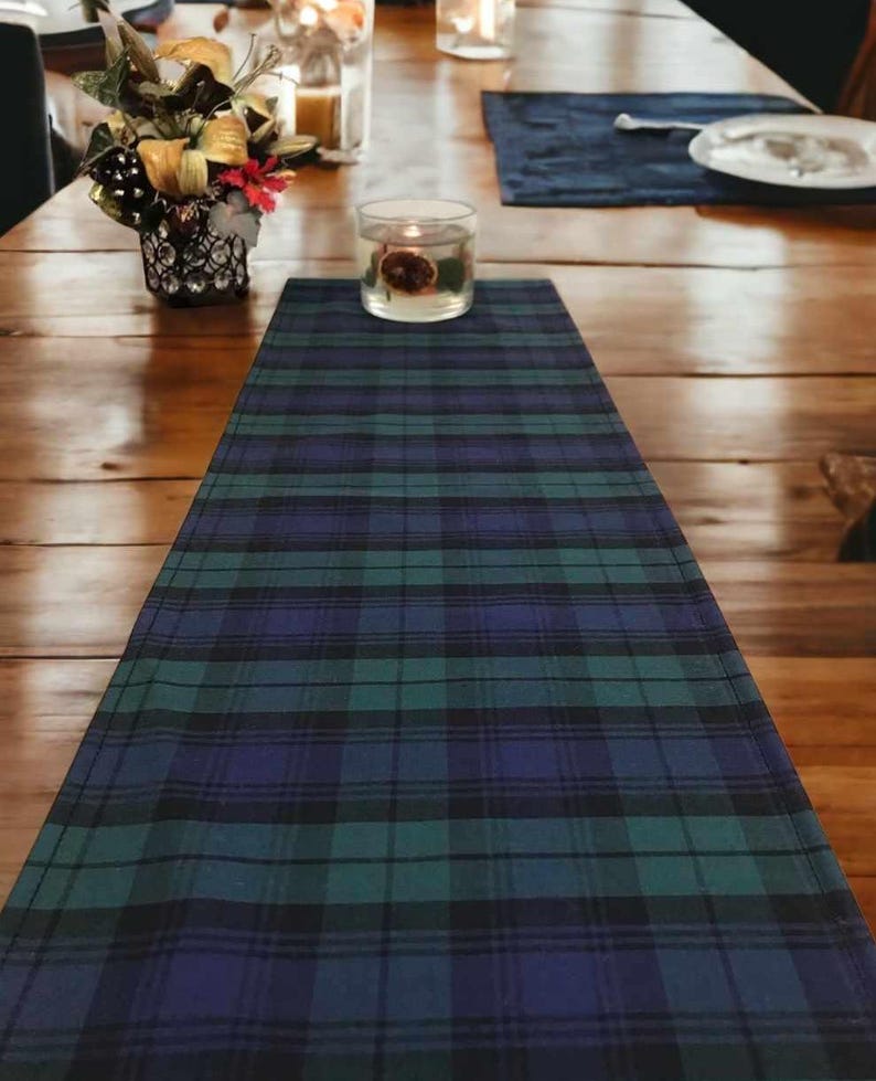 May include: A blue, green, and black plaid table runner. The runner is on a wooden table with a candle in a glass jar.