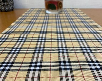 May include: A beige table runner with a black, white, and red plaid pattern. The runner is on a wooden table with a clear glass candle holder in the background.