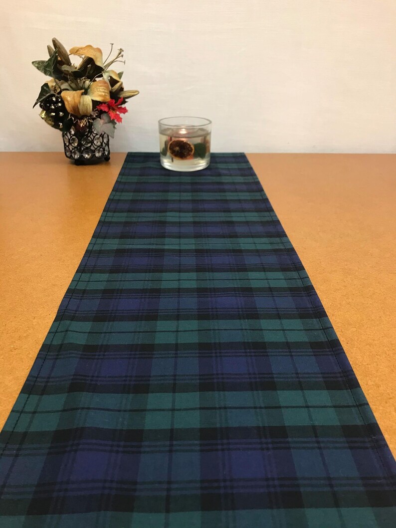 May include: A green, blue, and black plaid table runner. The runner is on a brown table with a clear glass candle holder in the center.