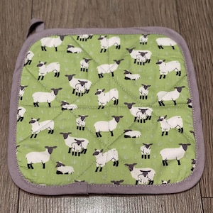 Op de afbeelding: Een groene, gestikte ovenwant met een patroon van witte schapen. De schapen hebben zwarte gezichten en een wit lammetje ernaast.