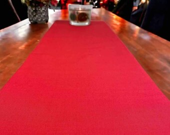 May include: A vibrant red table runner is displayed on a wooden table, creating a festive atmosphere. A small candle in a glass holder and a decorative vase with flowers are also on the table. The background features a Christmas tree.