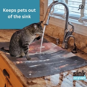 May include: A gray tabby cat drinks water from a faucet in a kitchen. The faucet is chrome and the countertop is granite. A mat with a dark abstract design is on the counter. Text on the image reads "Keeps pets out of the sink" and "sink out of sight."