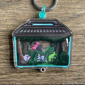 Könnte beinhalten: Ein durchsichtiger Acryl-Schlüsselanhänger mit einem schwarzen und grauen Mülleimer-Design. Im Inneren des Mülleimers befinden sich vier kleine, bunte Kreaturen in einer grünen Waldlandschaft.