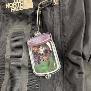 Könnte beinhalten: Ein klarer Acryl-Anhänger mit einer skurrilen Waldszene in einem Glas, mit einem lila Deckel. Der Anhänger ist an einem schwarzen Rucksack mit einem silbernen Verschluss befestigt. Das The North Face-Logo ist sichtbar.