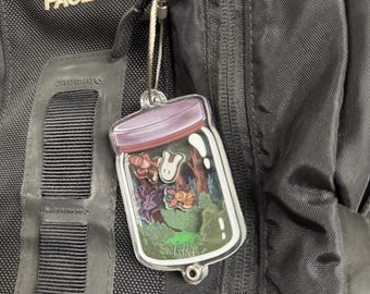 May include: A clear acrylic charm featuring a whimsical forest scene inside a jar, with a purple lid. The charm is attached to a black backpack with a silver clasp. The North Face logo is visible on the backpack.