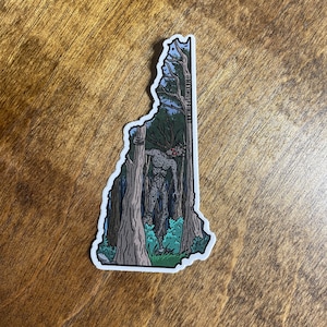 May include: A sticker of the state of New Hampshire with a creature from the woods with red eyes and a dark gray body. The creature is standing in a forest with tall trees and green foliage.