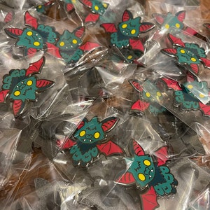 May include: A close-up of a pile of individually packaged green and red enamel pins. Each pin features a cartoon bat with yellow eyes and a toothy grin.