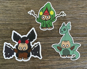 May include: Three cartoon character stickers with a green, brown, and black color scheme. The characters are wearing costumes of a leafy plant, a bat, and a dragon.