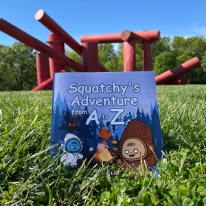 May include: A blue book with the title "Squatchy's Adventure from A to Z" on a green grassy field with a red metal sculpture in the background.